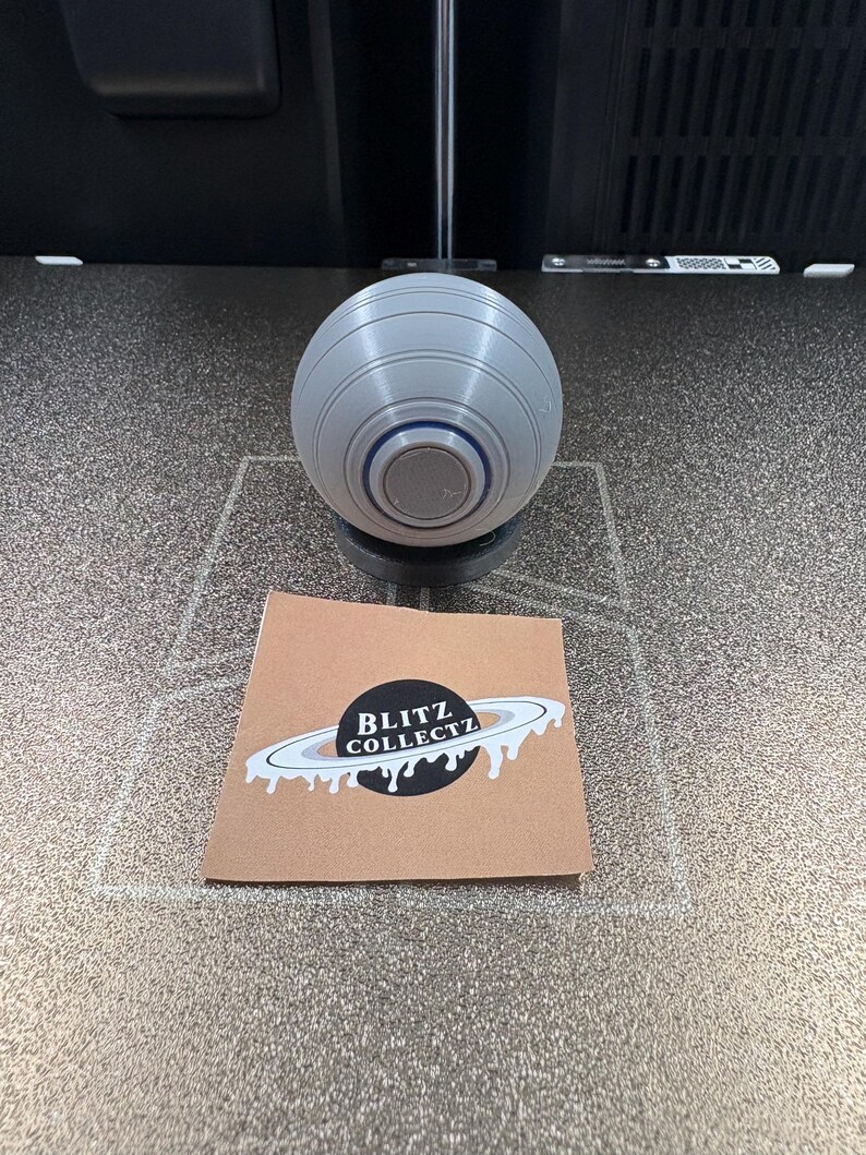 May include: A gray, spherical object with a circular base, featuring concentric rings and a central button. A small, brown card with the text "BLITZ COLLECTZ" and a planet graphic is also present.