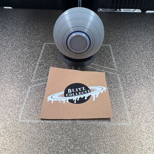 May include: A gray, spherical object with concentric rings and a dark center, resting on a black base. A brown card with the text "BLITZ COLLECTZ" and a planet design is in the foreground.