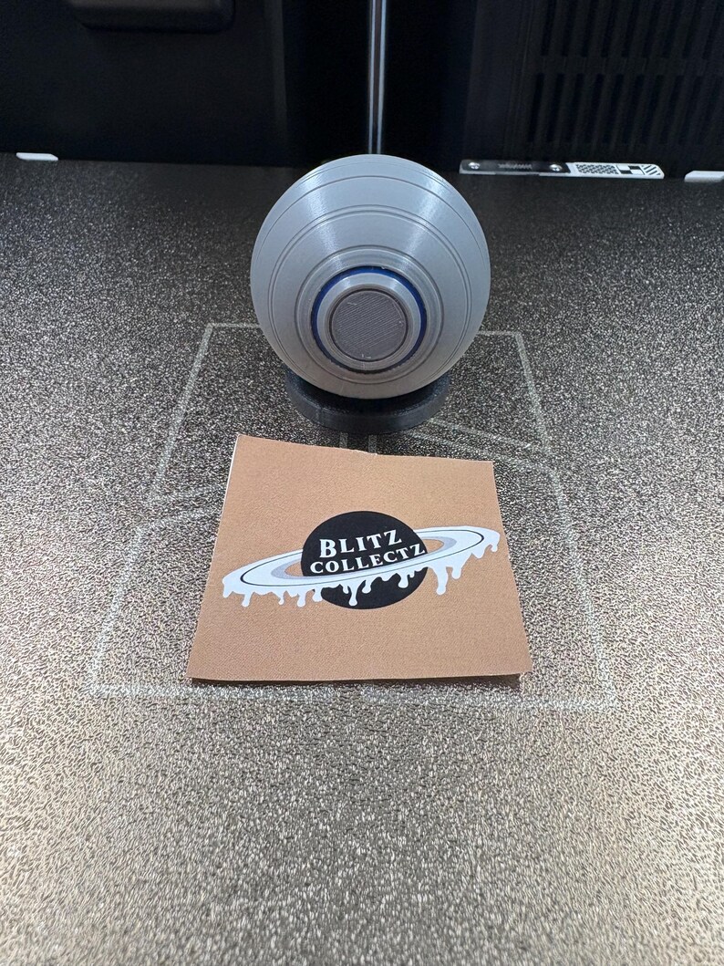 May include: A gray, spherical object with concentric rings and a dark center, resting on a black base. A brown card with the text "BLITZ COLLECTZ" and a planet design is in the foreground.