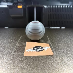 May include: A gray, spherical object with horizontal lines and a red accent sits on a surface. A small sticker with the text "BLITZ COLLECTZ" and a planet design is also present.