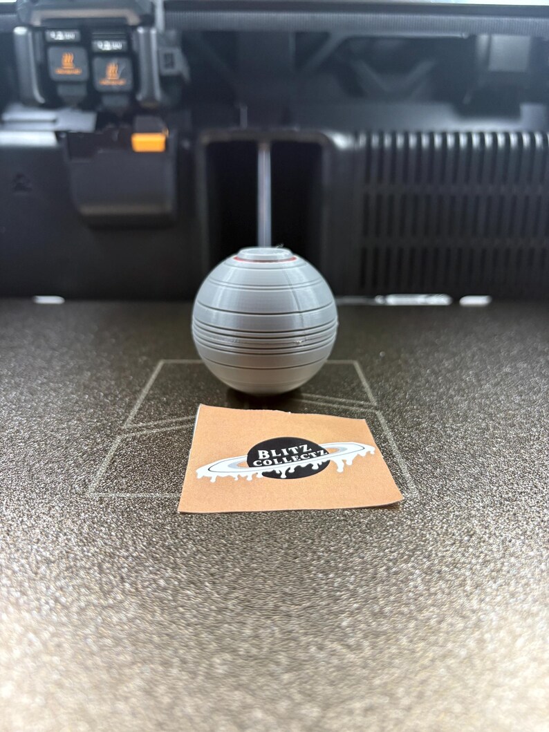 May include: A gray, spherical object with horizontal lines and a red accent sits on a surface. A small sticker with the text "BLITZ COLLECTZ" and a planet design is also present.