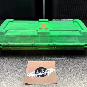 May include: A green rectangular storage container with orange arrow symbols on the lid. The container has a textured surface and is closed. A small brown label with the text "BLITZ COLLECTZ" is in the foreground.