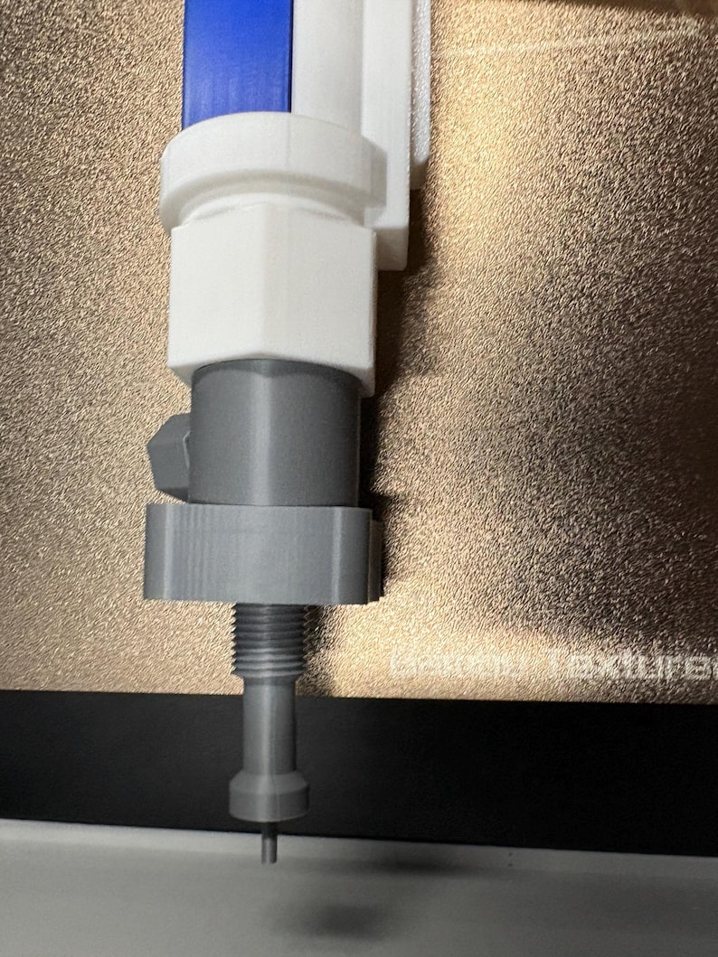 Op de afbeelding: Een 3D-geprint object in blauw, wit en grijs. Het heeft een cilindrisch ontwerp met een schroefdraad aan de onderkant en een fijne punt. Het object lijkt een mechanisch onderdeel te zijn, mogelijk voor een machine.
