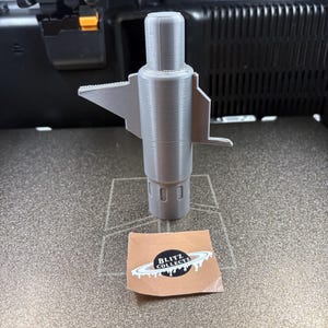 K&ouml;nnte beinhalten: Ein graues 3D-gedrucktes Objekt mit zylindrischem K&ouml;rper und fl&uuml;gelartigen Ausbuchtungen. Das Objekt steht auf einer strukturierten Oberfl&auml;che, neben einem Aufkleber mit dem Text "BLITZ COLLECTZ".