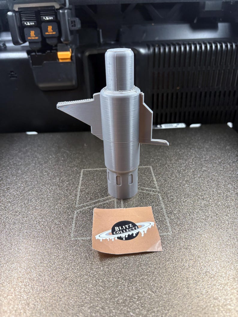 K&ouml;nnte beinhalten: Ein graues 3D-gedrucktes Objekt mit zylindrischem K&ouml;rper und fl&uuml;gelartigen Ausbuchtungen. Das Objekt steht auf einer strukturierten Oberfl&auml;che, neben einem Aufkleber mit dem Text "BLITZ COLLECTZ".