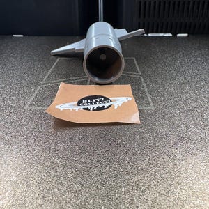 K&ouml;nnte beinhalten: Ein graues, 3D-gedrucktes Modell eines Strahltriebwerks mit Fl&uuml;geln. Ein Aufkleber mit dem Text "BLITZ COLLECTZ" und einem Planeten-Design befindet sich im Vordergrund. Das Modell befindet sich auf einer strukturierten Oberfl&auml;che.