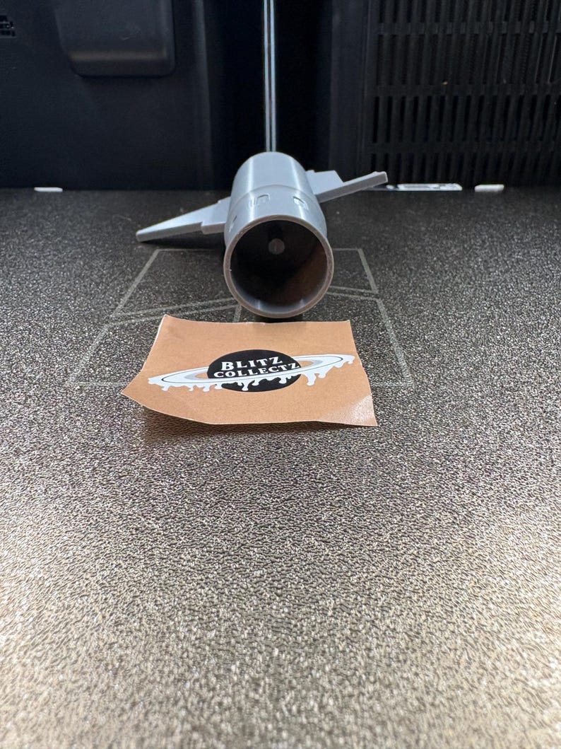 K&ouml;nnte beinhalten: Ein graues, 3D-gedrucktes Modell eines Strahltriebwerks mit Fl&uuml;geln. Ein Aufkleber mit dem Text "BLITZ COLLECTZ" und einem Planeten-Design befindet sich im Vordergrund. Das Modell befindet sich auf einer strukturierten Oberfl&auml;che.