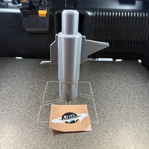 K&ouml;nnte beinhalten: Ein graues 3D-gedrucktes Objekt mit zylindrischem K&ouml;rper und fl&uuml;gelartigen Ausbuchtungen. Auf einer strukturierten Oberfl&auml;che befindet sich ein Aufkleber mit der Aufschrift "BLITZ COLLECTZ". Es scheint ein Prototyp oder eine Komponente zu sein.