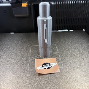 K&ouml;nnte beinhalten: Ein graues, 3D-gedrucktes zylindrisches Objekt, m&ouml;glicherweise ein Stift oder Werkzeug, mit einem Griff. Es steht auf einer strukturierten Oberfl&auml;che. Ein kleiner Aufkleber mit "BLITZ COLLECT" und einem Planeten-Design befindet sich daneben.