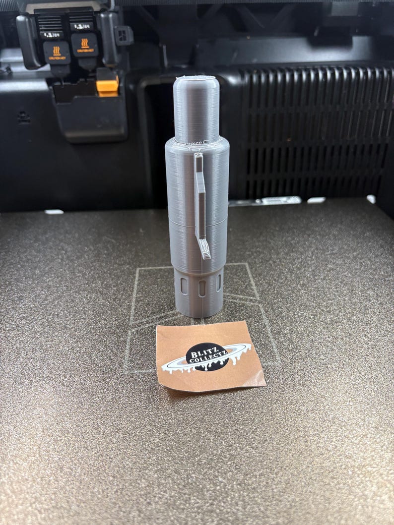 K&ouml;nnte beinhalten: Ein graues, 3D-gedrucktes zylindrisches Objekt, m&ouml;glicherweise ein Stift oder Werkzeug, mit einem Griff. Es steht auf einer strukturierten Oberfl&auml;che. Ein kleiner Aufkleber mit "BLITZ COLLECT" und einem Planeten-Design befindet sich daneben.
