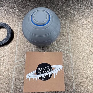 May include: A gray, spherical object with a blue ring and a circular top. A black and white sticker with the text "BLITZ COLLECTZ" and a planet design is also present. A black ring is also visible.