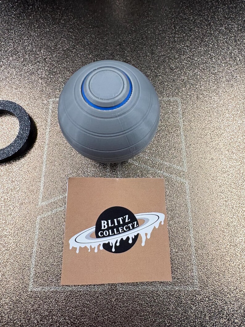 May include: A gray, spherical object with a blue ring and a circular top. A black and white sticker with the text "BLITZ COLLECTZ" and a planet design is also present. A black ring is also visible.
