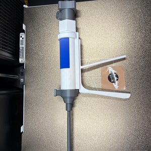 Op de afbeelding: Een 3D-geprint apparaat in wit en grijs, met een blauw accent, met een lange, slanke grijze staaf. Het apparaat heeft een handvat en een mondstukachtige bovenkant. Een sticker met tekst is zichtbaar aan de rechterkant.