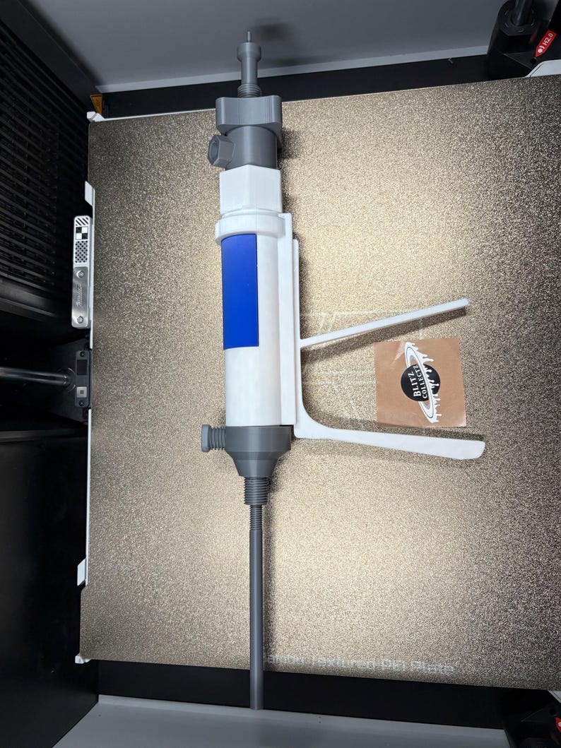 Op de afbeelding: Een 3D-geprint apparaat in wit en grijs, met een blauw accent, met een lange, slanke grijze staaf. Het apparaat heeft een handvat en een mondstukachtige bovenkant. Een sticker met tekst is zichtbaar aan de rechterkant.