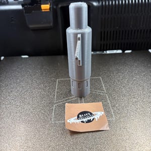 K&ouml;nnte beinhalten: Ein graues, zylindrisches 3D-gedrucktes Objekt mit detailliertem Design, das ein griffartiges Element und eine Reihe kreisf&ouml;rmiger Rillen aufweist. Ein kleiner Aufkleber mit dem Text "BLITZ COLLECTZ" ist ebenfalls vorhanden.