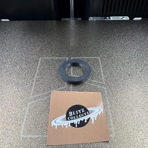 May include: A black, circular object with a hole in the center sits on a textured surface. A small, square sticker with the text "BLITZ COLLECTZ" and a planet design is also present.