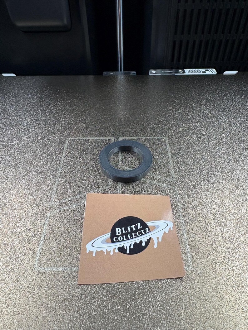 May include: A black, circular object with a hole in the center sits on a textured surface. A small, square sticker with the text "BLITZ COLLECTZ" and a planet design is also present.