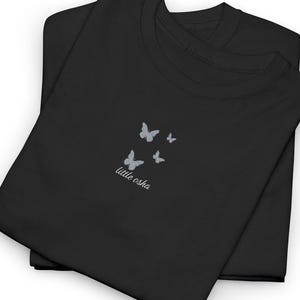 Può includere: Due magliette nere sono piegate e impilate. La maglietta superiore presenta un piccolo disegno di quattro farfalle azzurre, con la scritta "little osha" sotto. Le magliette sono realizzate in un materiale morbido.