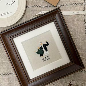 May include: A dark brown framed embroidery of two figures, with the initials "LJ & PL" and the date "2025.8.30". The artwork is set against a white mat. The background includes a ribbon and a card. The frame is dark brown.