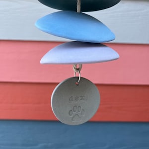 Może przedstawiać: Szary ceramiczny wiatraczek z odciskiem łapy i wygrawerowanym imieniem "dex". Wiatraczek składa się z czterech ceramicznych dysk&oacute;w w odcieniach niebieskiego i fioletowego, wiszących na sznurku.