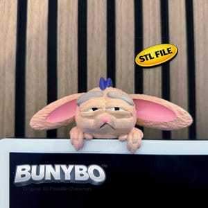 May include: A 3D-printed character with a grumpy expression, featuring large pink-lined ears and a blue tuft of hair. The character is perched on a white surface, with the "BUNYBO" logo visible below. A yellow oval sticker reads "STL FILE".