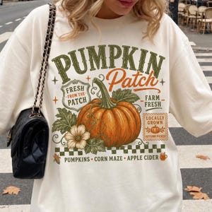 May include: An off-white crewneck sweatshirt featuring a large graphic print of an orange pumpkin with green leaves and vines. The text reads PUMPKIN Patch, FRESH FROM THE PATCH, FARM FRESH, LOCALLY GROWN, AUTUMN PICKED, PUMPKINS, CORN MAZE, APPLE CIDER.
