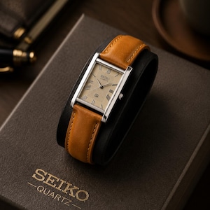 Pode incluir: Um relógio Seiko Quartz retangular com caixa prateada e pulseira de couro castanho claro. O mostrador do relógio é bege claro com algarismos romanos. O relógio é exibido em um suporte preto, em cima de uma caixa castanha com o logotipo Seiko Quartz.