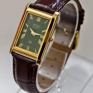 Pode incluir: Um relógio Seiko Quartz retangular dourado com mostrador verde e pulseira de couro castanho. O mostrador do relógio apresenta algarismos romanos e ponteiros dourados. A pulseira é de couro castanho escuro texturizado. O relógio é exibido em um suporte transparente.
