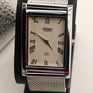 May include: A rectangular Seiko Quartz watch with a silver mesh band. The watch face is off-white with black Roman numerals and hands. The watch is displayed on a black stand.