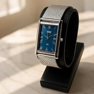 Może przedstawiać: Prostokątny zegarek Seiko Quartz z niebieską tarczą i srebrną bransoletą z siatki. Zegarek posiada rzymskie cyfry i jest prezentowany na czarnym stojaku. Tarcza zegarka ma szerokość około 2,5 cm.