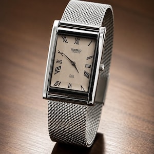 Könnte beinhalten: Eine rechteckige Seiko Quartz-Uhr mit einem silbernen Mesh-Armband. Das Zifferblatt der Uhr ist cremefarben mit schwarzen römischen Ziffern und Zeigern. Die Uhr wird auf einer Holzoberfläche präsentiert.