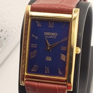 May include: A gold-toned Seiko Quartz watch with a rectangular blue face and Roman numeral hour markers. The watch has a brown leather band. The watch face displays the Seiko logo and the word "QUARTZ".