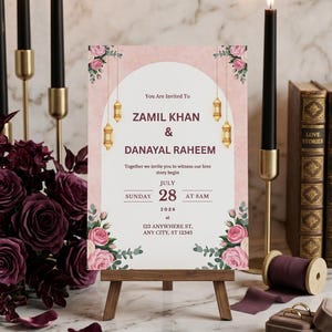 Puede incluir: Una tarjeta de invitaci&oacute;n de boda sobre un caballete de madera con un arco blanco, fondo rosa texturizado y detalles florales. El texto dice: You Are Invited To ZAMIL KHAN & DANAYAL RAHEEM, Together we invite you to witness our love story begin, JULY SUNDAY 28 AT 8AM 2026 at 123 ANYWHERE ST, ANY CITY, ST 12345.