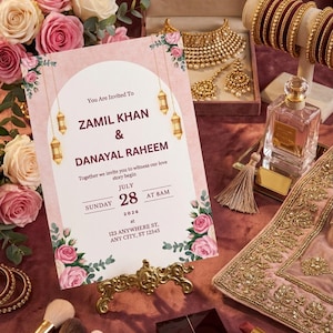 Puede incluir: Una tarjeta de invitaci&oacute;n de boda con un arco blanco, linternas doradas colgantes y rosas rosadas. El texto dice Zamil Khan y Danayal Raheem, domingo 28 de julio de 2026 a las 8 a.m., 123 Anywhere St, Any City, ST 12345, sobre un soporte dorado.