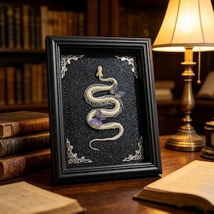 May include: A framed artwork featuring a snake skeleton and two purple butterflies on a black textured background. The black frame has decorative silver corner accents. The artwork is displayed on a wooden surface.