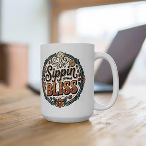 May include: A white ceramic mug featuring a circular graphic with the text Sippin BLISS in a stylized font. The design includes orange slices, coffee beans, and floral motifs in shades of orange, green, and black, surrounded by a dark border.