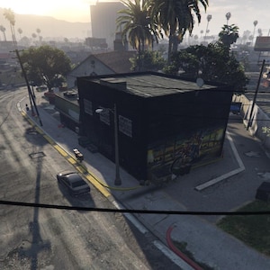 Può includere: Vista aerea di una strada cittadina con un edificio nero al centro. La strada si snoda attraverso la scena, con linee elettriche e palme. Un'auto è parcheggiata sul lato della strada.