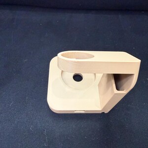 Puede incluir: Un soporte para tel&eacute;fono beige impreso en 3D con un soporte circular, un peque&ntilde;o compartimento rectangular y un agujero central. El soporte tiene una base cuadrada redondeada y est&aacute; dise&ntilde;ado para sostener un tel&eacute;fono.