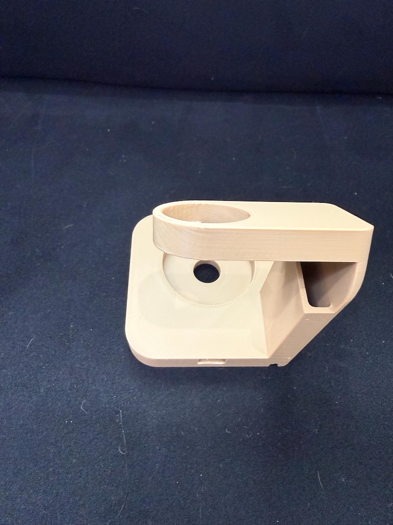 Puede incluir: Un soporte para tel&eacute;fono beige impreso en 3D con un soporte circular, un peque&ntilde;o compartimento rectangular y un agujero central. El soporte tiene una base cuadrada redondeada y est&aacute; dise&ntilde;ado para sostener un tel&eacute;fono.