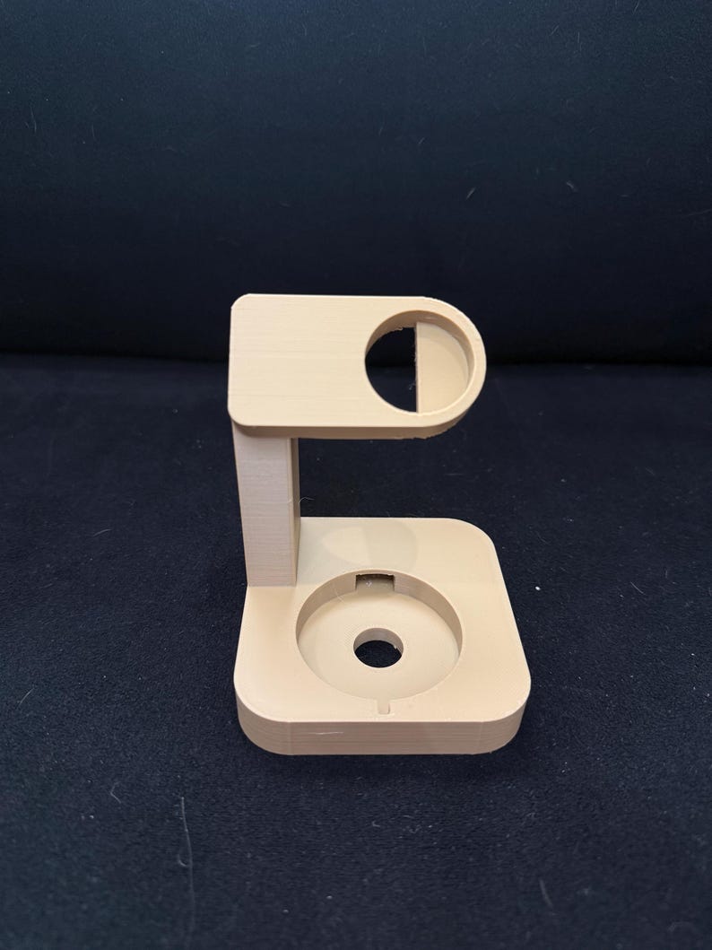 Puede incluir: Un soporte beige impreso en 3D con una parte superior e inferior circulares, conectado por un soporte vertical. La parte superior presenta un recorte semicircular, y la base tiene una hendidura circular central. El soporte est&aacute; dise&ntilde;ado para sostener un art&iacute;culo.