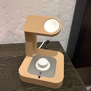 Puede incluir: Un soporte de carga de color beige para un reloj inteligente. El soporte tiene una almohadilla de carga circular y una base cuadrada con un &aacute;rea de carga plateada. Se ve un disco de carga blanco en la parte superior.