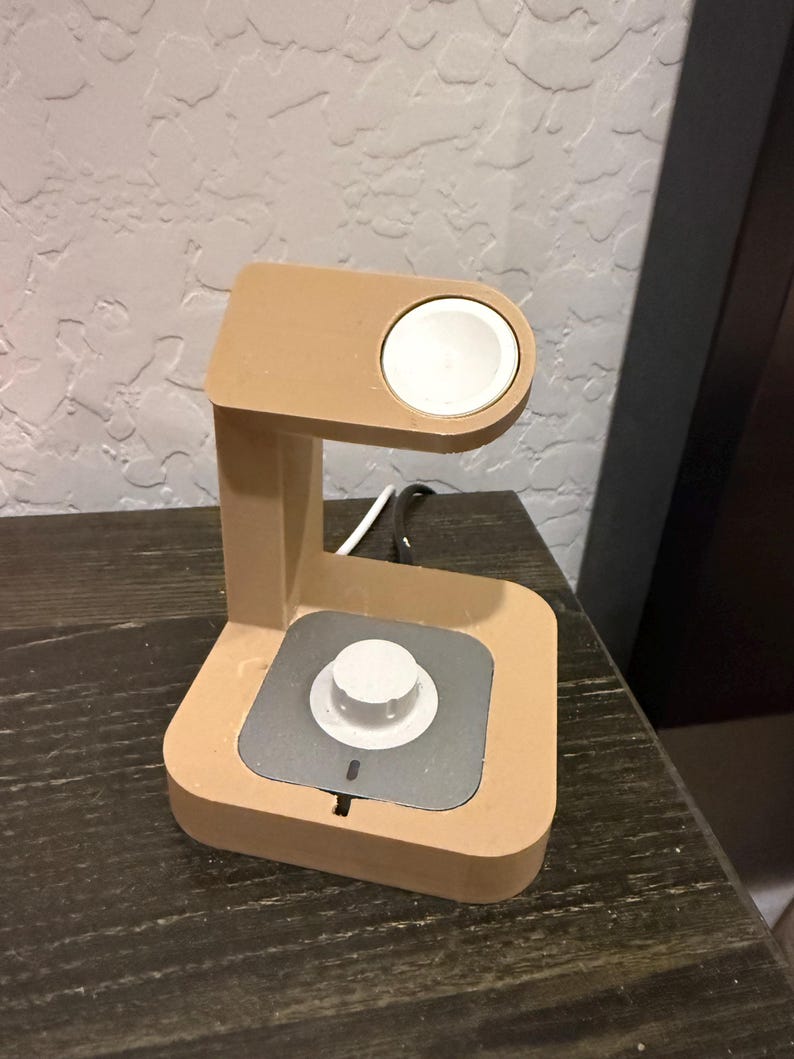 Puede incluir: Un soporte de carga de color beige para un reloj inteligente. El soporte tiene una almohadilla de carga circular y una base cuadrada con un &aacute;rea de carga plateada. Se ve un disco de carga blanco en la parte superior.