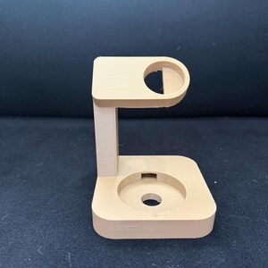 Puede incluir: Un soporte de reloj beige impreso en 3D. El soporte tiene una base rectangular con una hendidura circular y un soporte vertical que conduce a una parte superior curva con una abertura circular. El soporte est&aacute; dise&ntilde;ado para sostener un reloj inteligente.