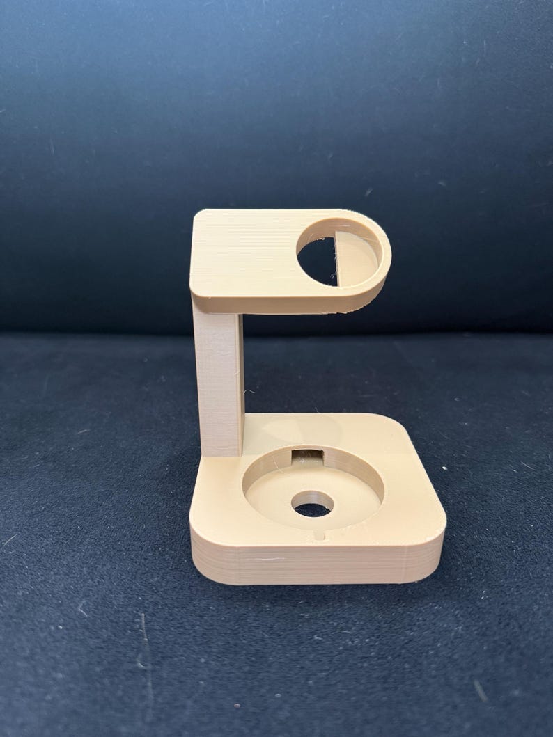 Puede incluir: Un soporte de reloj beige impreso en 3D. El soporte tiene una base rectangular con una hendidura circular y un soporte vertical que conduce a una parte superior curva con una abertura circular. El soporte est&aacute; dise&ntilde;ado para sostener un reloj inteligente.