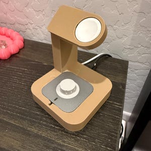 Puede incluir: Un soporte de carga de color beige para smartwatch. El soporte tiene una plataforma de carga circular y una base cuadrada con un centro plateado. Un cable de carga blanco est&aacute; conectado al soporte.