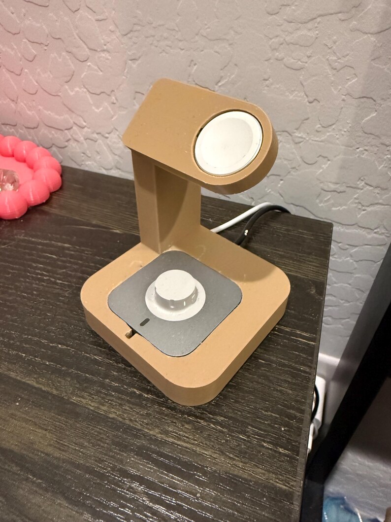Puede incluir: Un soporte de carga de color beige para smartwatch. El soporte tiene una plataforma de carga circular y una base cuadrada con un centro plateado. Un cable de carga blanco est&aacute; conectado al soporte.