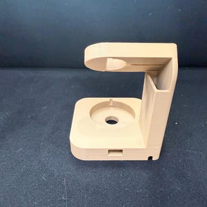 Puede incluir: Un soporte beige impreso en 3D con una base circular y una secci&oacute;n superior curvada. El soporte tiene una ranura rectangular y un agujero cil&iacute;ndrico. El dise&ntilde;o parece ser para sujetar un dispositivo electr&oacute;nico.