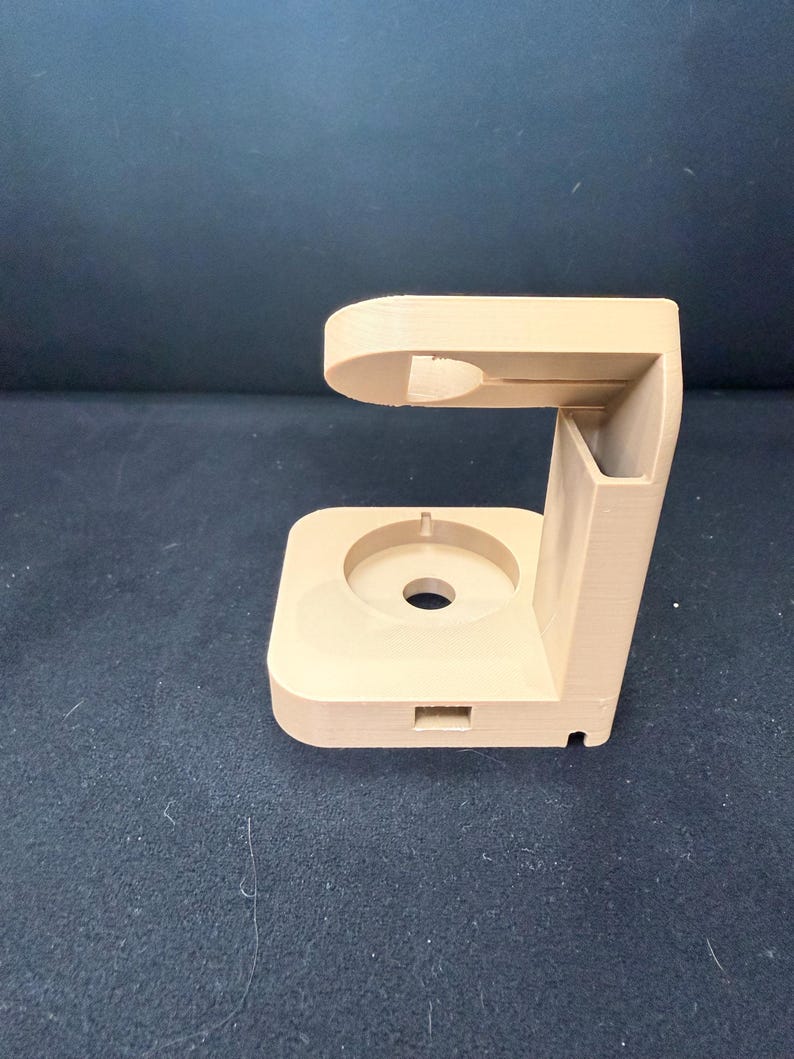 Puede incluir: Un soporte beige impreso en 3D con una base circular y una secci&oacute;n superior curvada. El soporte tiene una ranura rectangular y un agujero cil&iacute;ndrico. El dise&ntilde;o parece ser para sujetar un dispositivo electr&oacute;nico.