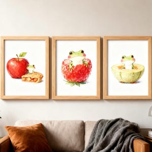以下が含まれることがあります： 3つの額入り水彩画プリント。気まぐれなカエルのイラストが特徴です。リンゴとパイを持つカエル、イチゴの中のカエル、メロンの中のカエルが描かれています。フレームは薄茶色で、アートワークは白い背景に配置されています。
