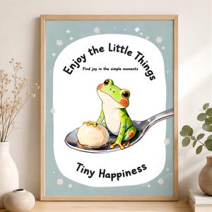 May include: A framed wall art print featuring a green frog sitting on a silver spoon next to a white round object. The text reads: Enjoy the Little Things, Find joy in the simple moments, and Tiny Happiness, set against a light blue background.